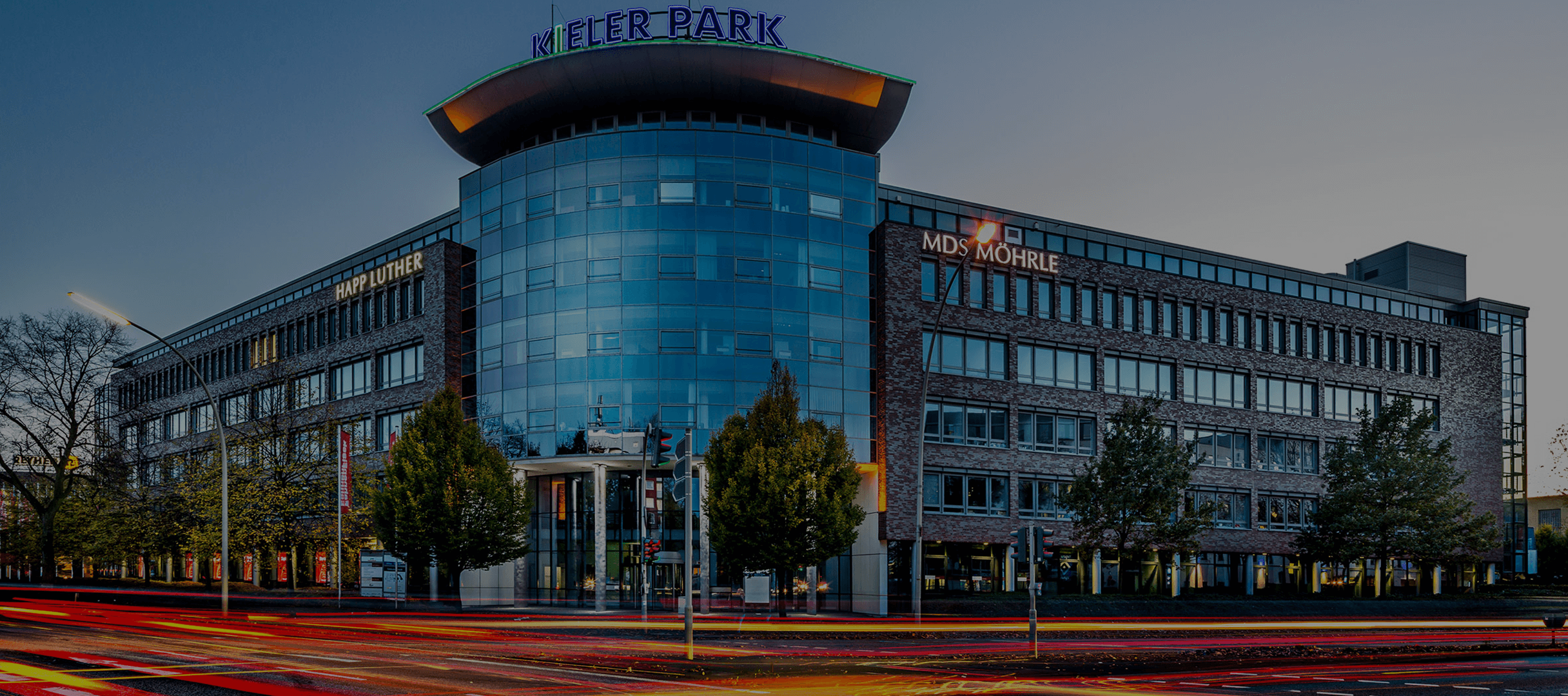 Ein modernes Gebäude mit Glasfassade und Backsteinfassade, das in der Abenddämmerung beleuchtet wird. Es trägt die Schilder „Meier Park“ und „MDS Möhrle“. Im Vordergrund sind Lichtspuren vorbeifahrender Fahrzeuge zu sehen. Bäume säumen die Straße.