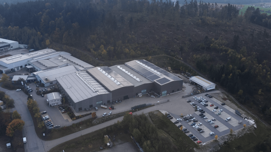 Luftaufnahme eines Industriekomplexes in einer Waldlandschaft. Die Gebäude haben graue Dächer und sind von Parkplätzen mit Fahrzeugen umgeben. Im Hintergrund ist ein gerodeter Baumbestand zu sehen.