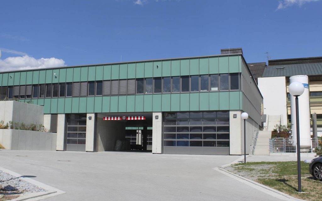 Ein modernes, zweistöckiges Industriegebäude mit grüner Fassade und großen Glasfenstern. Es verfügt über zwei Garagentore und ist von einem gepflasterten Bereich mit Straßenlaternen umgeben. Der Himmel ist klar mit ein paar Wolken.