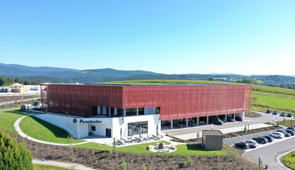 Ein modernes Gebäude mit einer roten Netzfassade in einer grünen Landschaft mit Hügeln und Bäumen im Hintergrund. Das Gebäude ist von einem Parkplatz und Wegen umgeben und überblickt einen klaren blauen Himmel.