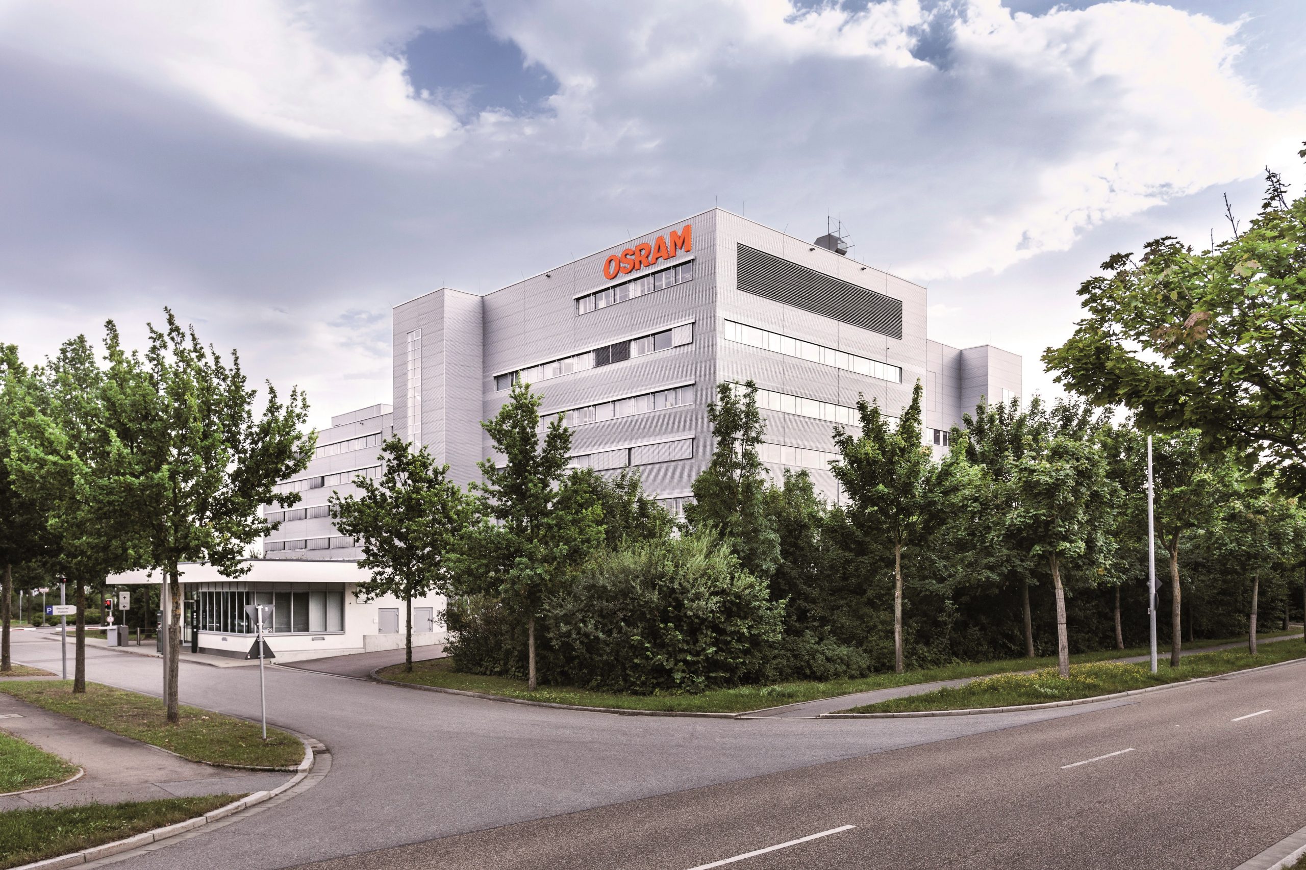 Modernes Bürogebäude mit weißer Fassade, umgeben von üppigen grünen Bäumen und einem sauberen Bürgersteig. Das Gebäude hat mehrere Stockwerke und trägt oben in orangefarbenen Buchstaben „OSRAM“. Den Hintergrund bildet ein grauer, wolkiger Himmel.