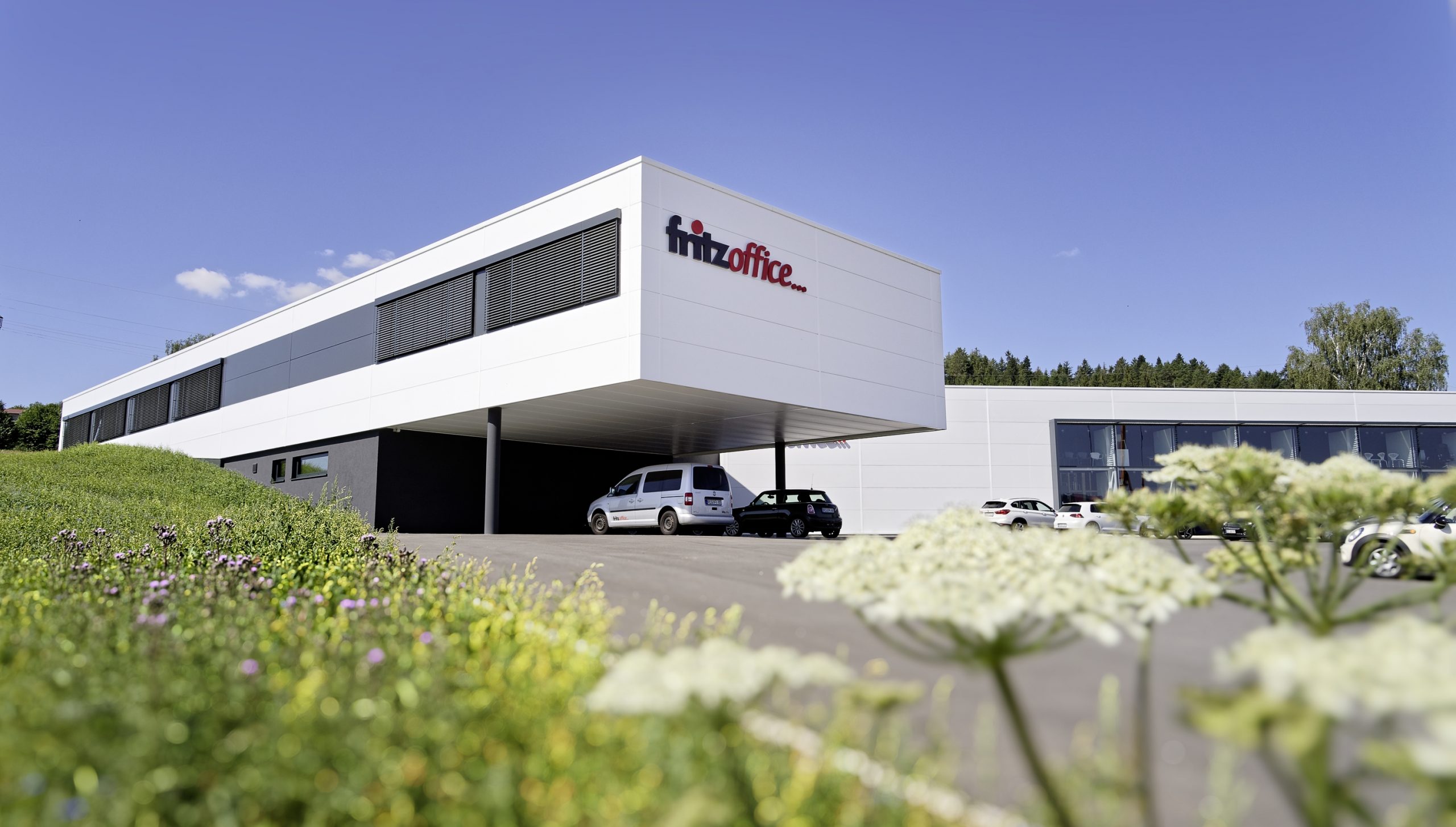 Modernes Bürogebäude mit einem Schild mit der Aufschrift „fitz office“ an der Fassade. Das Gebäude ist weiß und rechteckig und hat einen großen Glasbereich. Draußen parken mehrere Autos und im Vordergrund ist eine Grasfläche mit Wildblumen zu sehen.