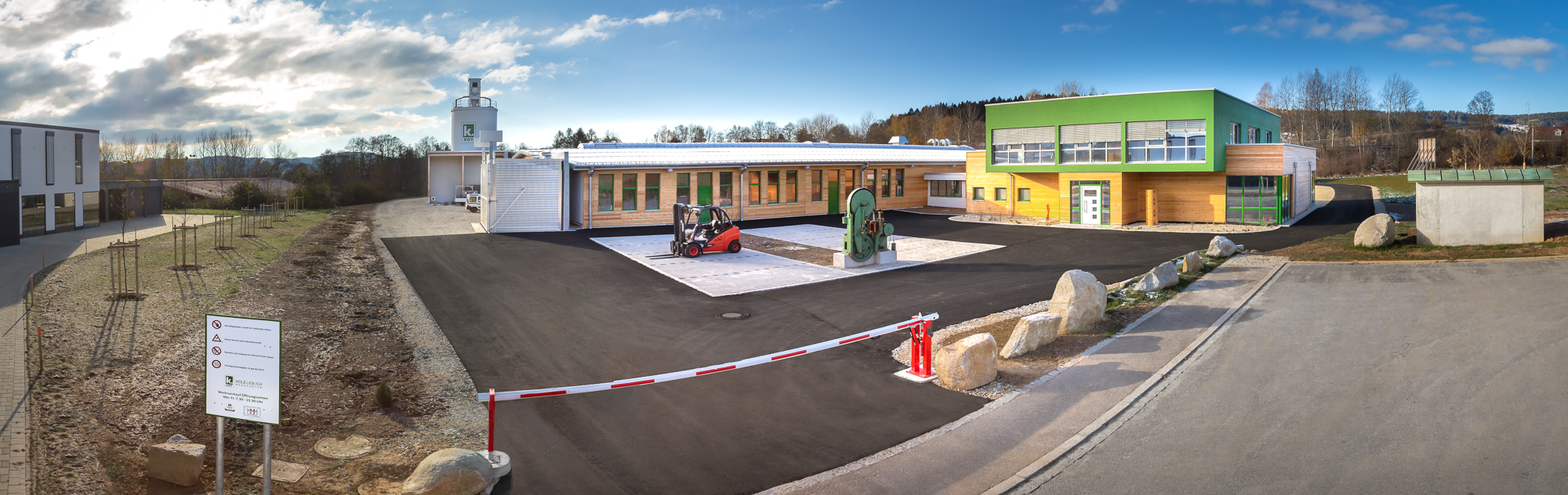 Panoramablick auf einen modernen Gebäudekomplex mit Grün- und Holzakzenten, umgeben von einer offenen Landschaft. Davor parkt ein kleiner Gabelstapler, im Vordergrund ist links ein Schrankenbaum mit Schildern zu sehen. Der Himmel ist teilweise bewölkt.