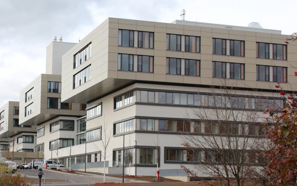 Modernes, mehrstöckiges Gebäude mit großen, kastenförmigen Abschnitten und vielen Fenstern. Die Fassade ist in neutralen Tönen gehalten. Im Vordergrund sind Bäume mit Herbstlaub zu sehen, im Hintergrund ist ein bewölkter Himmel zu sehen.