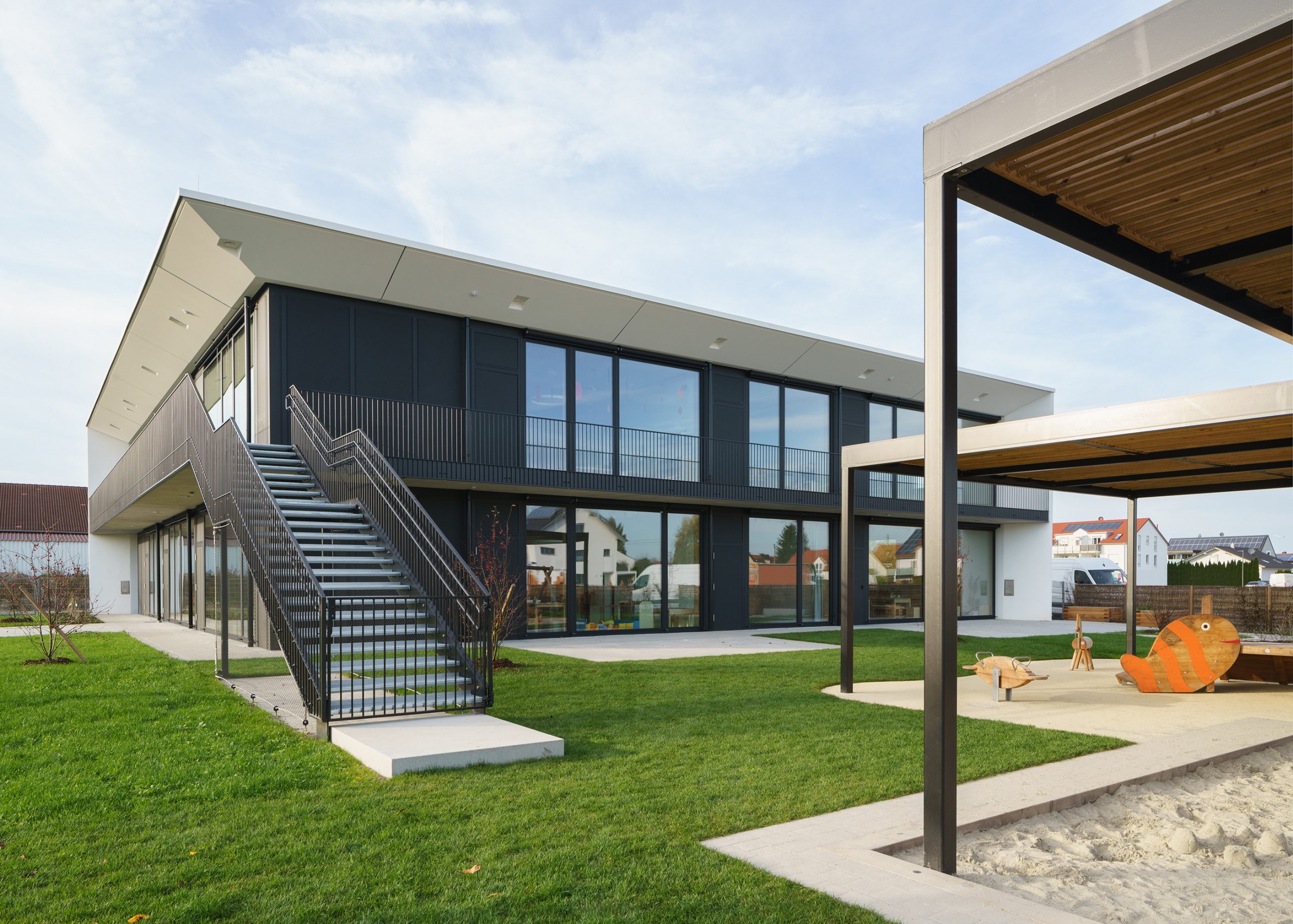 Ein modernes, zweistöckiges Gebäude mit großen Glasfenstern und einer Außentreppe. Die Umgebung besteht aus einem gepflegten Rasen und im Vordergrund ist ein überdachter Spielbereich mit einer farbenfrohen, fischförmigen Spielstruktur zu sehen.