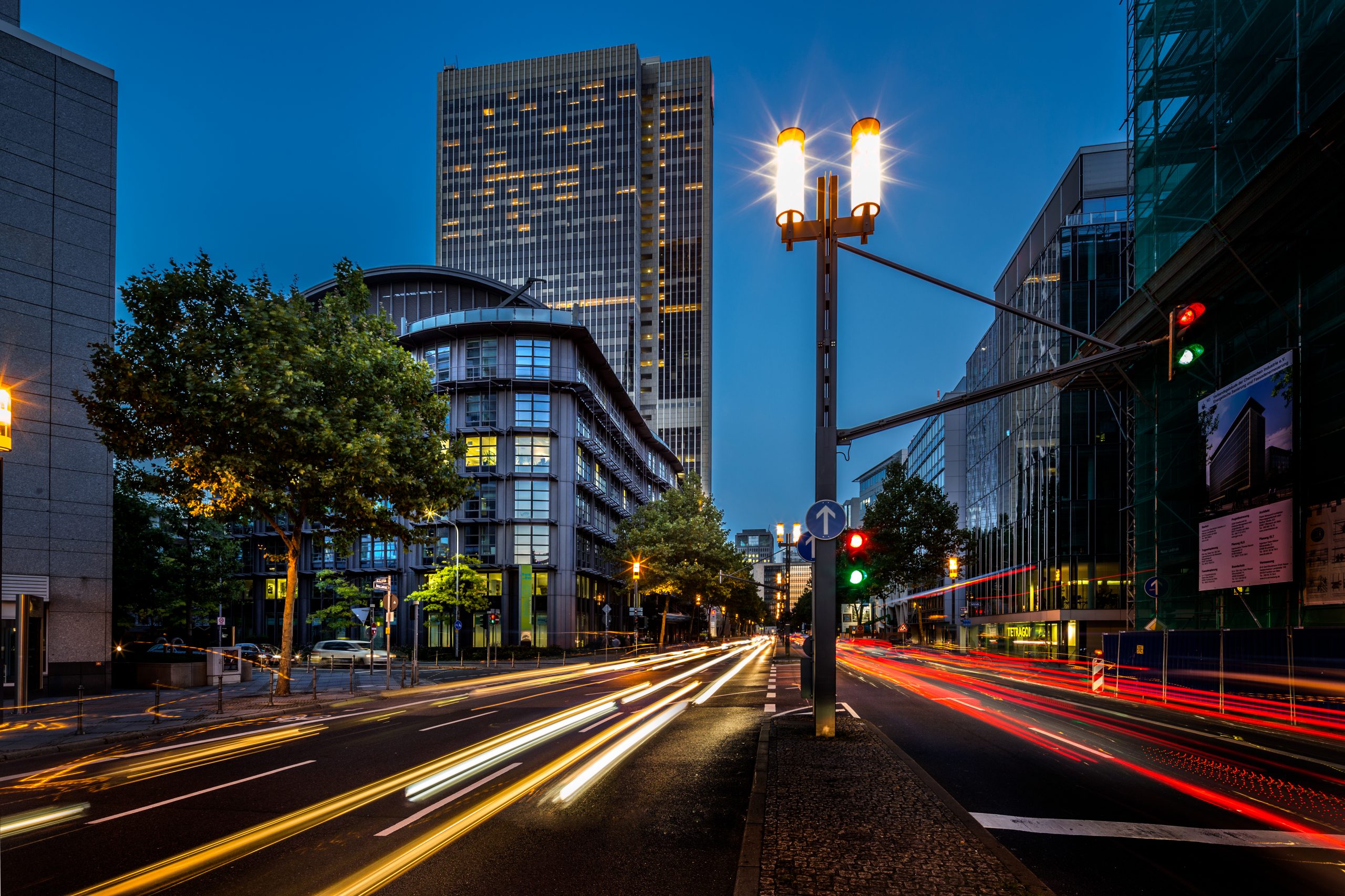 Eine Stadtstraße mit hohen Gebäuden in der Abenddämmerung. Lichtspuren von Fahrzeugen erzeugen dynamische Streifen auf der Straße. Der Himmel ist tiefblau und eine Straßenlaterne erhellt die Szene. Ampeln leuchten grün und rot.