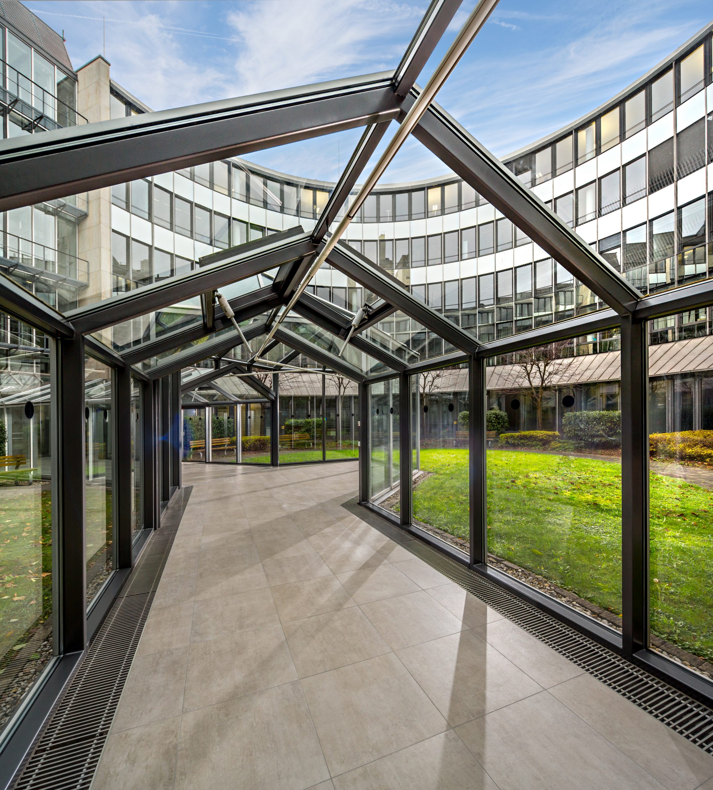 Ein moderner Glassteg mit Metallrahmen schlängelt sich durch einen Innenhof, der von einem runden Gebäude mit reflektierenden Fenstern umgeben ist. Der Innenhof ist mit grünem Gras und Sträuchern unter einem klaren blauen Himmel ausgestattet.