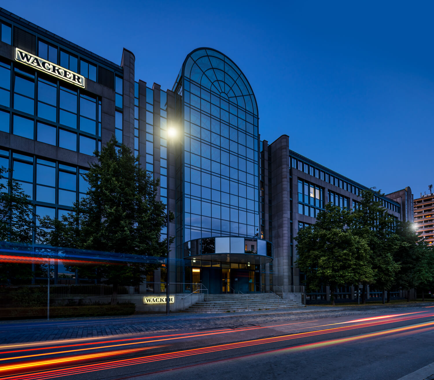 Ein modernes Bürogebäude mit großen Glasfenstern und einem gewölbten Eingang mit der Aufschrift „Wacker“. Die Szene ist in der Dämmerung beleuchtet, mit Lichtspuren vom vorbeifahrenden Verkehr auf der Straße davor. Bäume säumen den Bürgersteig.